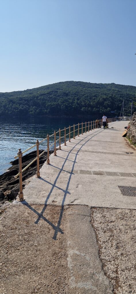 Popločana šetnica uz more vijuga uz rub vode, omeđena smeđom ogradom. Dvoje ljudi hoda u daljini, sa šumovitim brdom i mirnom plavom vodom pod vedrim nebom u pozadini.