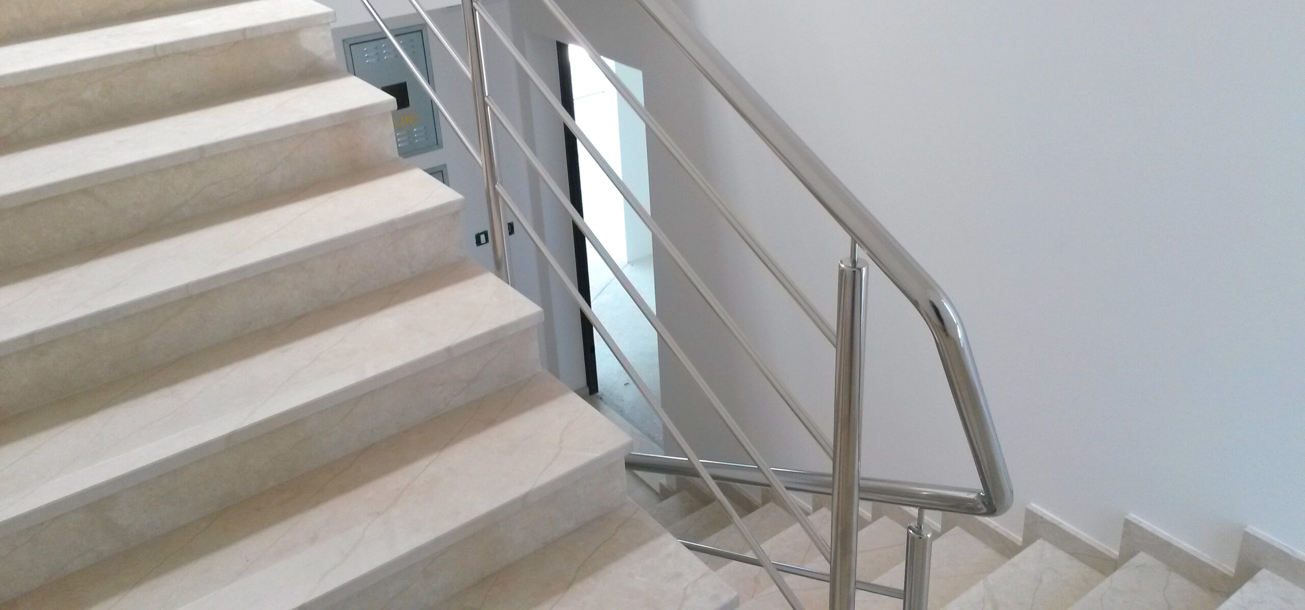 Stubište s bež mramornim stepenicama i modernom metalnom ogradom, koje vodi do malog odmorišta i vrata ispod, unutar čiste zgrade s bijelim zidovima.