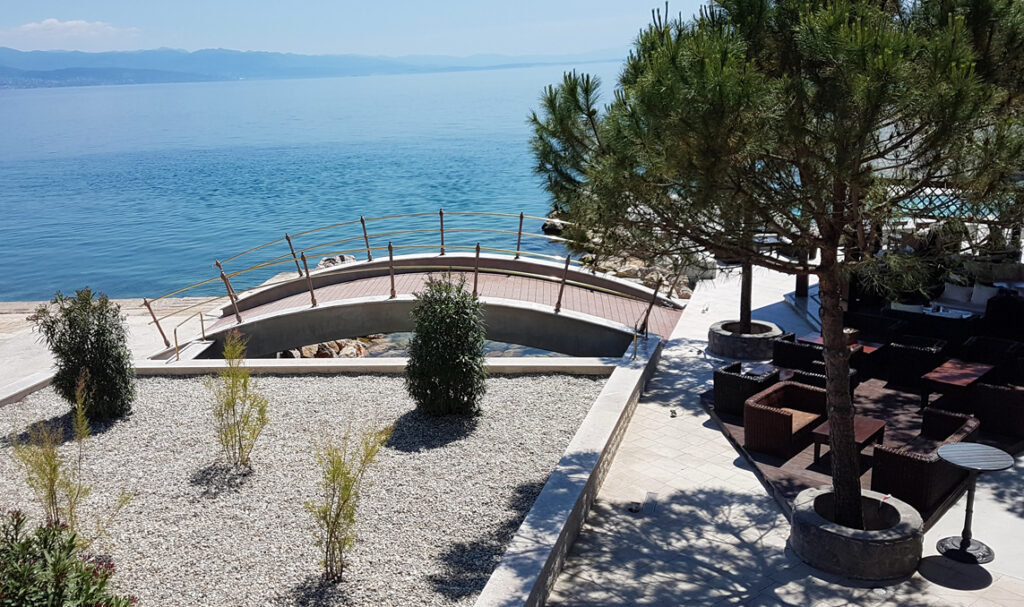 Zakrivljeni drveni most prelazi preko stjenovite obale u blizini mirnog plavog mora, s drvećem, grmljem i vanjskim sjedenjem pod vedrim, sunčanim nebom. Planine su vidljive u pozadini preko vode.