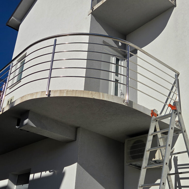 Metalne ljestve naslanjaju se na balkon moderne bijele zgrade pod vedrim plavim nebom. Balkon ima ograde od inoxa i mesinga, staklena vrata i klima uređaj postavljen odmah ispod.