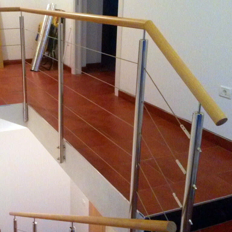 Moderna unutarnja ograda od inoxa s stupovima od nehrđajućeg čelika, horizontalnim čeličnim sajlama i svijetlim drvenim rukohvatom, postavljena uz stubište i hodnik s crvenim pločicama na podu.