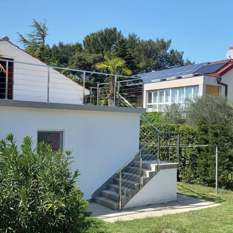 Moderna bijela zgrada s vanjskim stepenicama i ogradama od inoxa i mesinga, balkonom s palmom u tegli i solarnim panelima na obližnjem krovu. Zeleno grmlje i drveće okružuju područje pod vedrim plavim nebom.