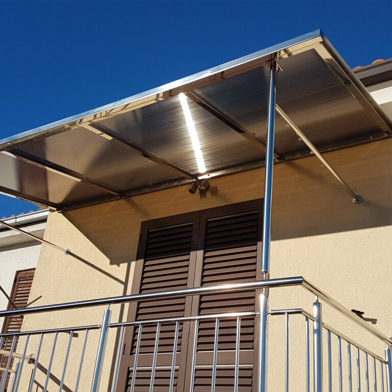 Moderni balkon s metalnom ogradom i prozirnom, kosom nadstrešnicom od inoxa koju podupiru stupovi od nehrđajućeg čelika, pričvršćenom iznad smeđih vrata s kapcima uz bež zid pod vedrim plavim nebom.