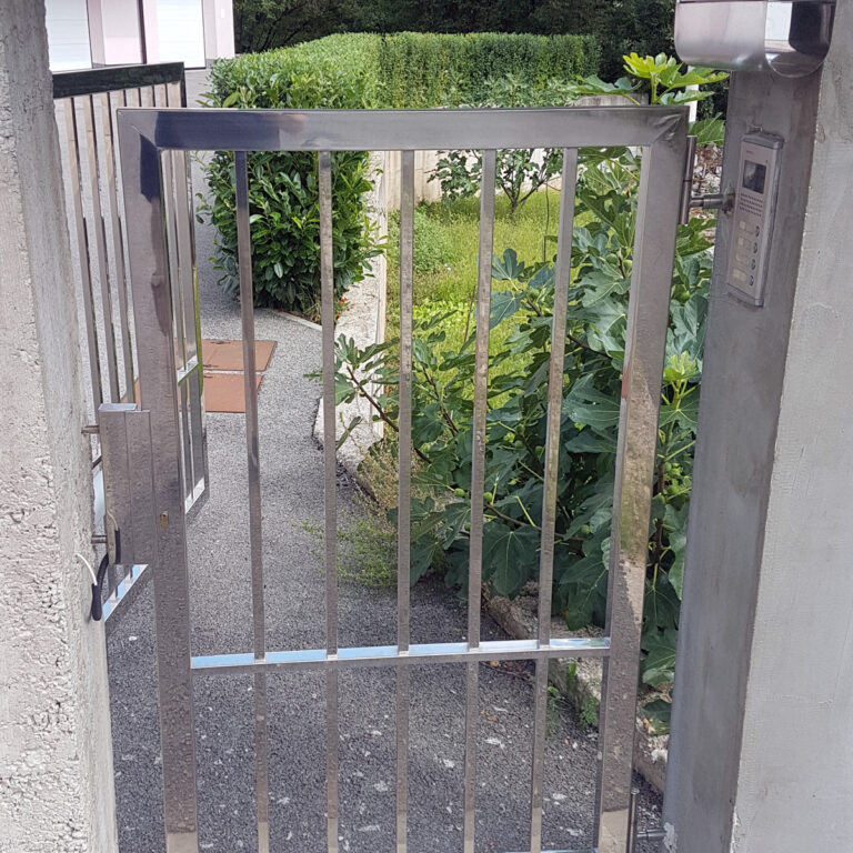 Sjajna portuni od inoxa vrata s okomitim šipkama pričvršćena su za betonski zid, djelomično otvoren, koji vodi u vrt sa zelenim grmljem i šljunčanom stazom. Interfon je postavljen na zidu pored vrata.