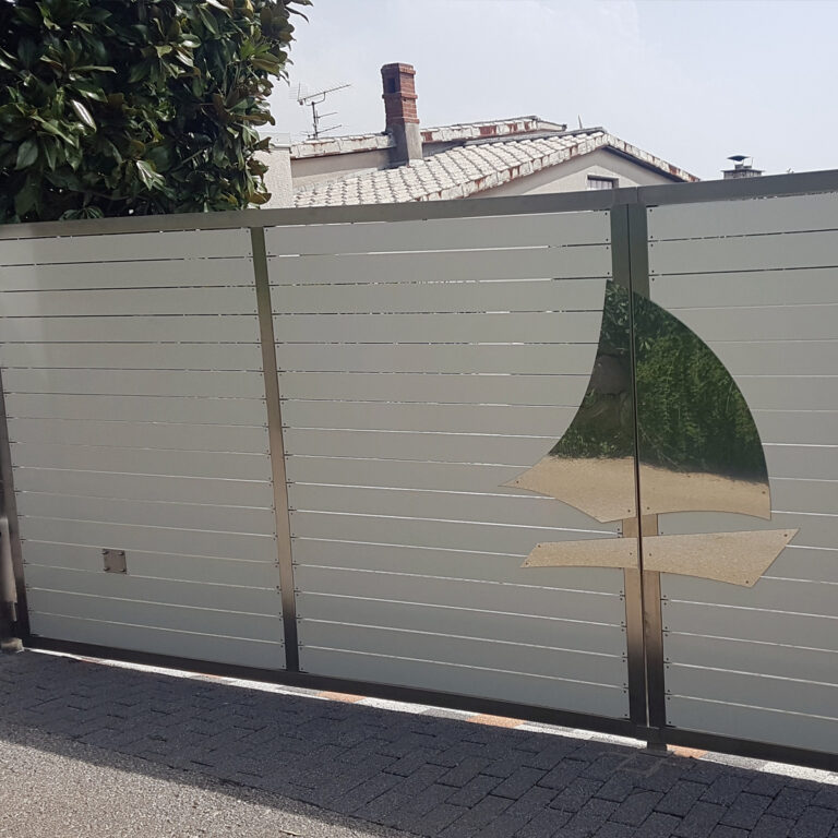 Moderna metalna vrata s horizontalnim letvicama i izrezanim dizajnom jedrilice, izrađena od inoxa materijala, nalaze se ispred kuće s crijepnim krovom i dimnjacima. Zeleno lišće vidljivo je s lijeve strane.