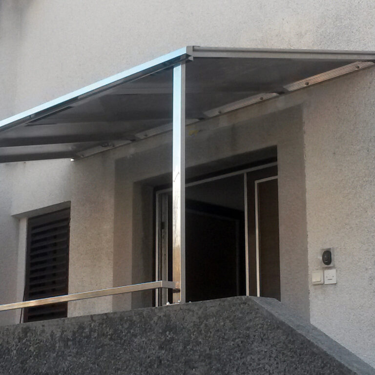 Moderna metalna i staklena tenda, ili nadstrešnice od inoxa, prekriva ulaz u sivu zgradu s kosom rampom i rukohvatom koji vode do otvorenih vrata. Prekidači za svjetlo ili zvona na vratima postavljeni su u blizini ulaza.