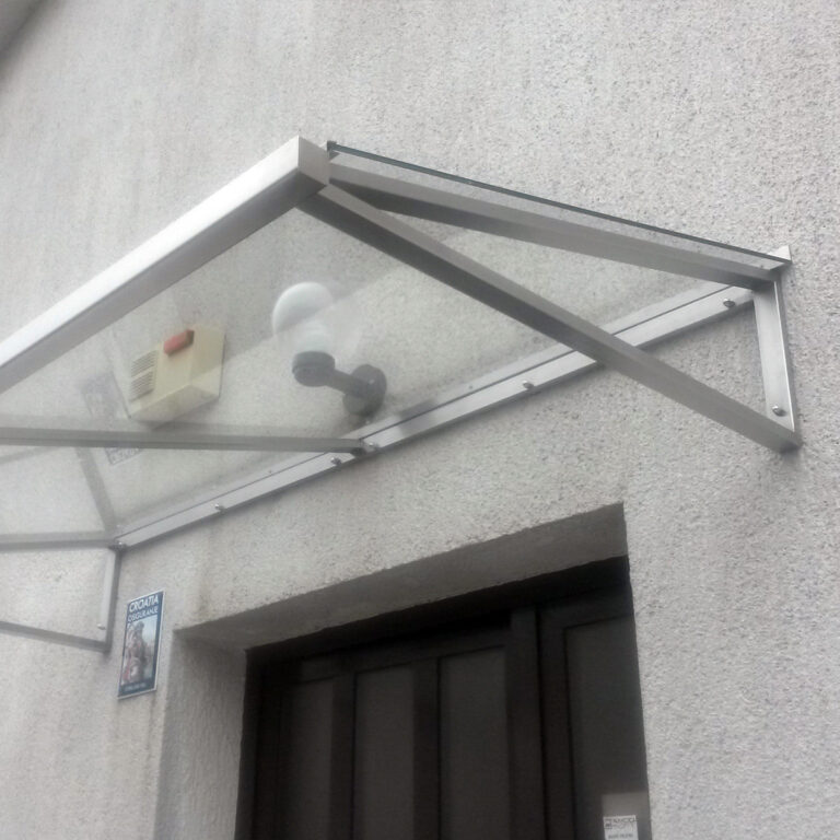 Staklena i metalna nadstrešnica od inoxa postavljena je iznad vrata na teksturiranom svijetlosivom zidu. Ispod tende nalaze se žarulja i sigurnosni uređaj, a na obližnjem zidu nalazi se mala obavijest.