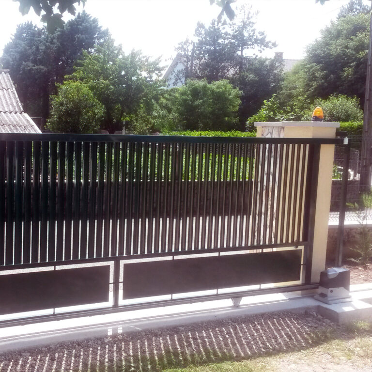 Crna metalna klizna vrata s okomitim šipkama, punim panelima na dnu i detaljima od inoxa djelomično su otvorena između dva kamena stupa. Drveće i kuće vidljivi su u pozadini za sunčanog dana.