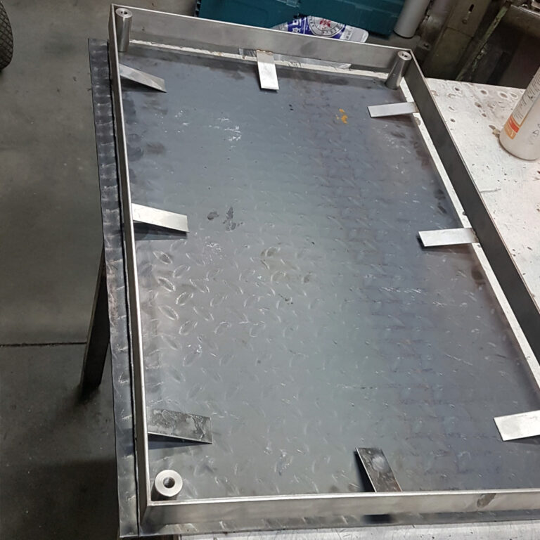 Pravokutni metalni lim od inox galanterije s malim metalnim nosačima zavarenim uz rubove, postavljen na radni stol u radioničkom okruženju. Alati i ostali materijali vidljivi su u pozadini.