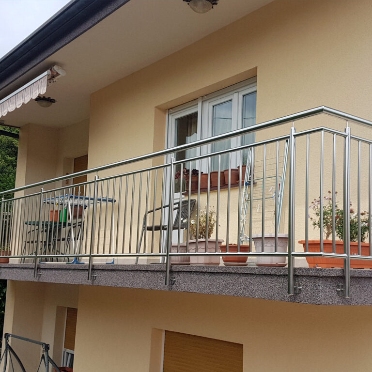 Balkon s ogradama od inoxa i mesinga, nekoliko biljaka u teglama, malim stolom i dvije stolice ispred bež kuće s velikim prozorom i tendom iznad.