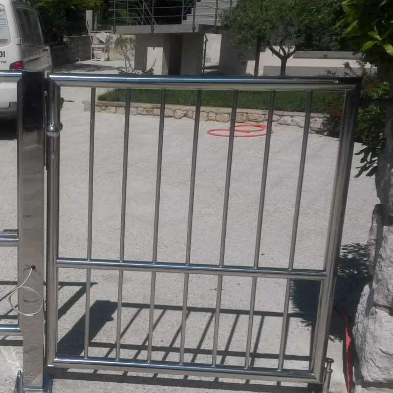 Sjajna vrata Portuni od inoxa s okomitim rešetkama zatvorena su preko prilaza. Iza njih, s lijeve strane parkiran je bijeli kombi, a u pozadini se vidi kuća sa stepenicama i zelenilom.