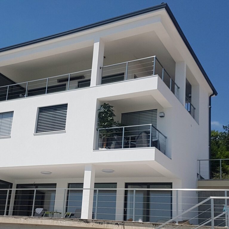 Moderna bijela kuća na tri kata s velikim balkonima, brojnim prozorima, biljkom i vanjskim namještajem na srednjem balkonu te vedrim plavim nebom u pozadini.