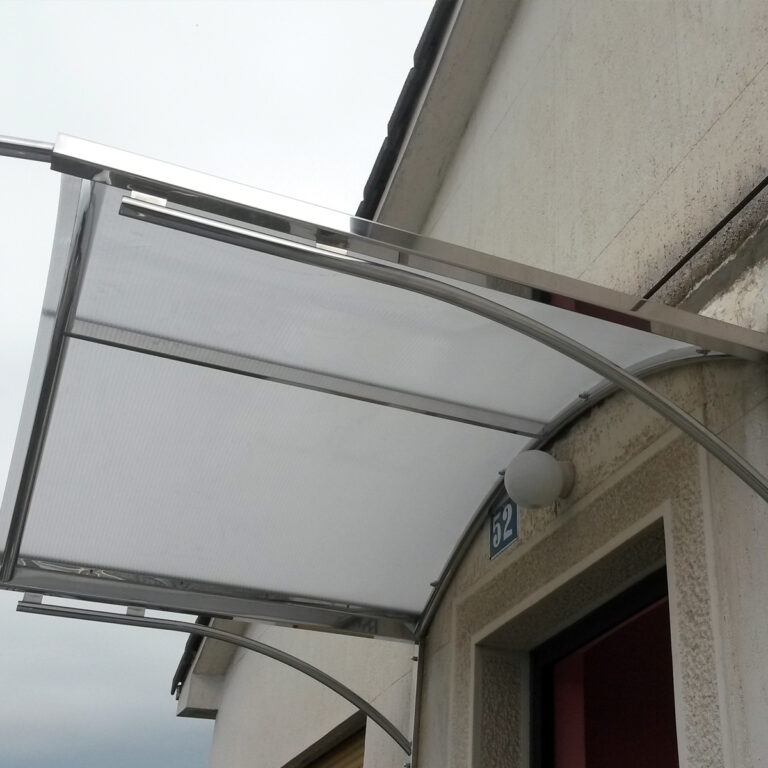 Moderna, zakrivljena nadstrešnica od inoxa i stakla postavljena je iznad vrata na svijetloj zgradi, nudeći zaklon i hlad. Zidna svjetiljka i broj adrese "52" vidljivi su u blizini vrata.