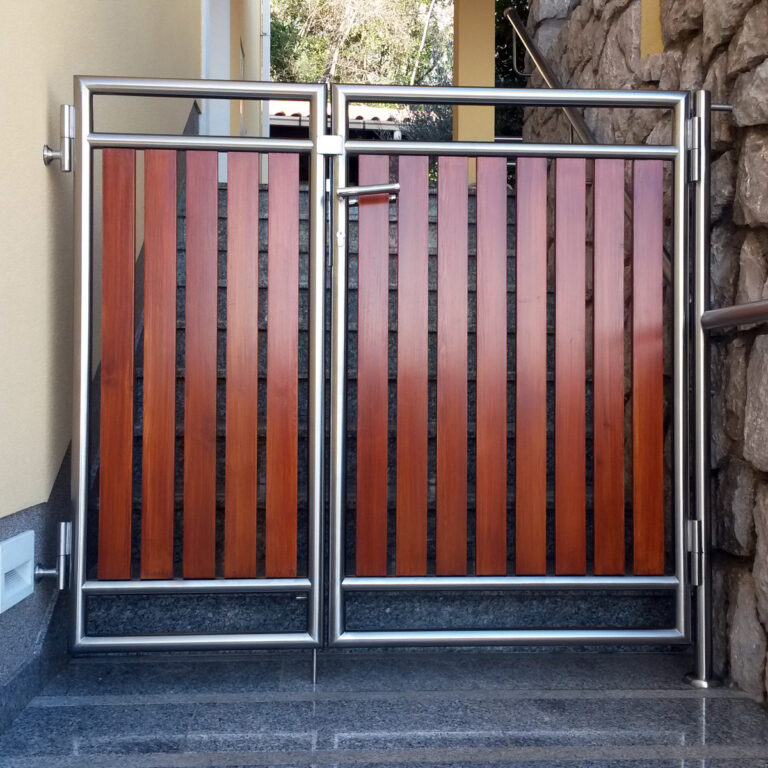 Moderna vrata od vertikalnih drvenih panela postavljena u okvir od inoxa, smještena na vrhu vanjskih kamenih stepenica pored kamenog zida i žute zgrade.