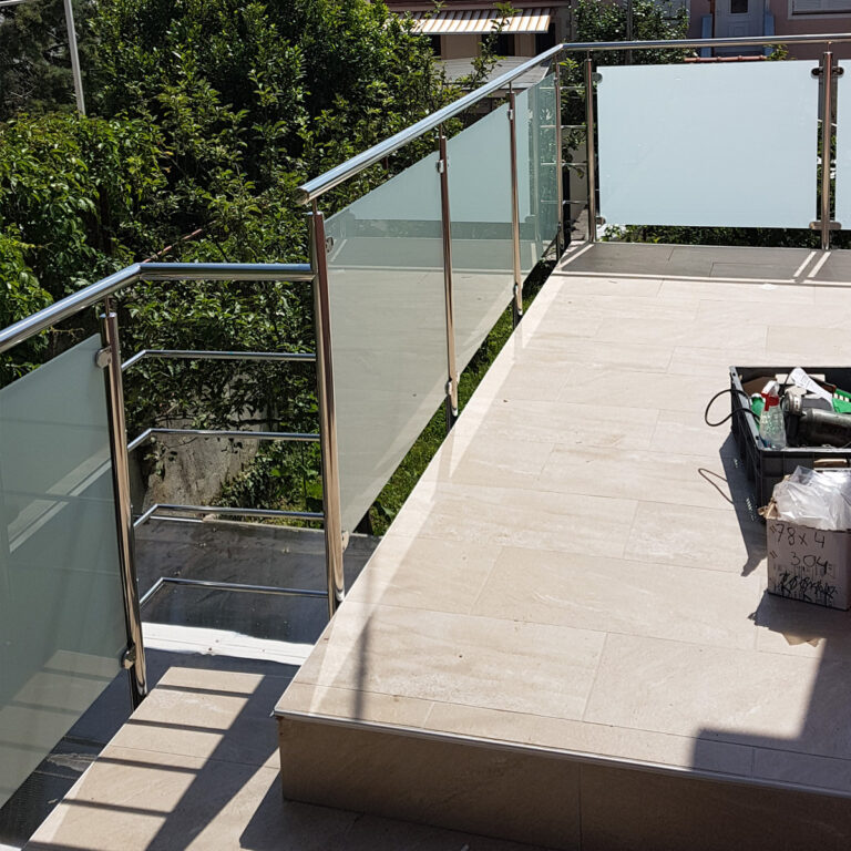 Moderni vanjski balkon ima ograde od inoxa i mesinga, bež pločice na podu i sunčevu svjetlost koja baca sjene. Kutija s alatom i materijali nalaze se na desnoj strani, dok zeleno drveće stvara mirnu kulisu.