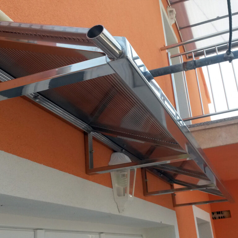 Krupni plan moderne nadstrešnice od inoxa s kosim prozirnim krovom, postavljene iznad vanjskih vrata na narančasto-bijeloj zgradi. Odvodna cijev proteže se s ruba tende.