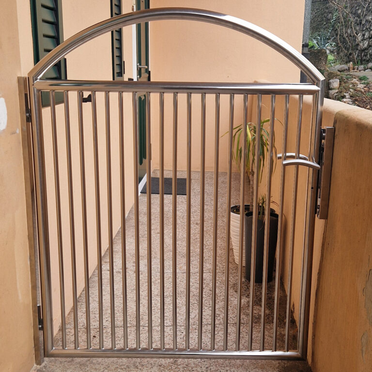 Metalna vrata s okomitim šipkama i lučnim vrhom, poznata kao portuni od inoxa, postavljena su na ulazu u prolaz pored bež zida. Saksija s biljkom nalazi se u kutu blizu vrata.
