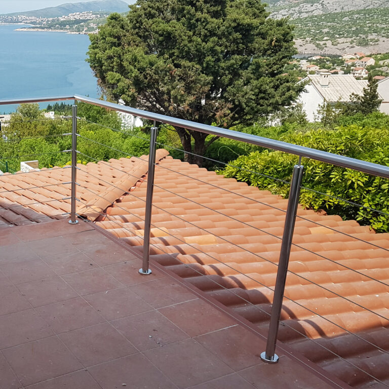 Terasa s ogradama od inoxa i mesinga pruža pogled na krov od crvenog crijepa, bujno zelenilo i vodenu površinu s udaljenim brdima i zgradama u pozadini.