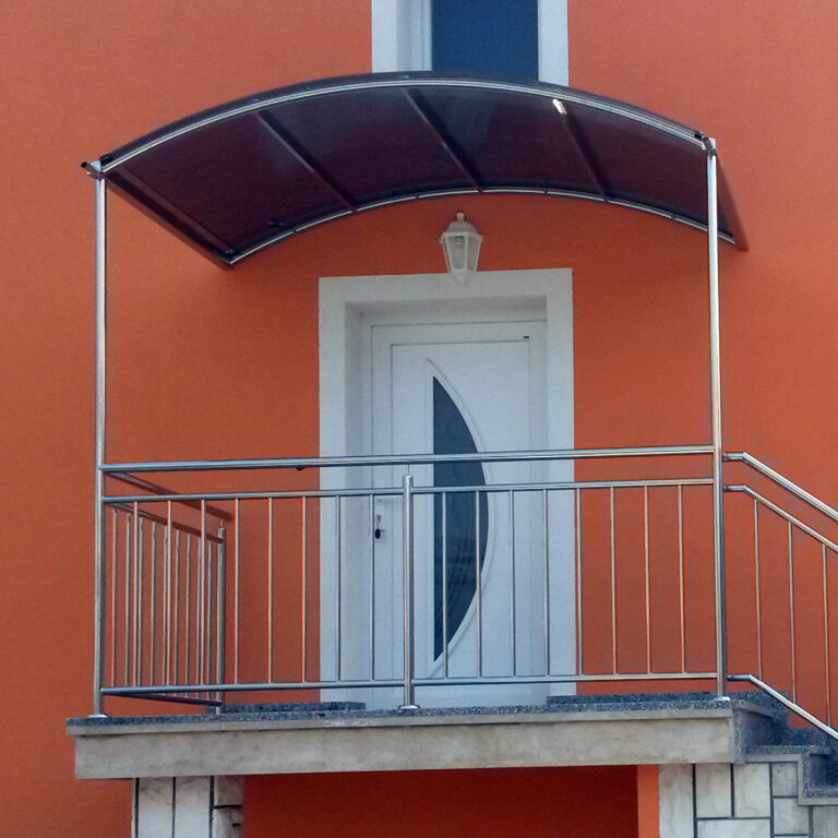 Bijela vrata sa zakrivljenim staklenim prozorom otvaraju se na mali balkon s metalnom ogradom i nadstrešnicom od inoxa, postavljenom uz narančasti vanjski zid. Iznad vrata visi lampa u obliku lampiona.
