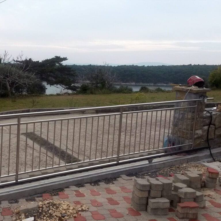 Popločano područje s metalnim vratima Portuni od inoxa u prvom planu, nekoliko naslaganih cigli i travnato dvorište iza njih. U daljini se pod oblačnim nebom vide drveće i rijeka ili jezero.