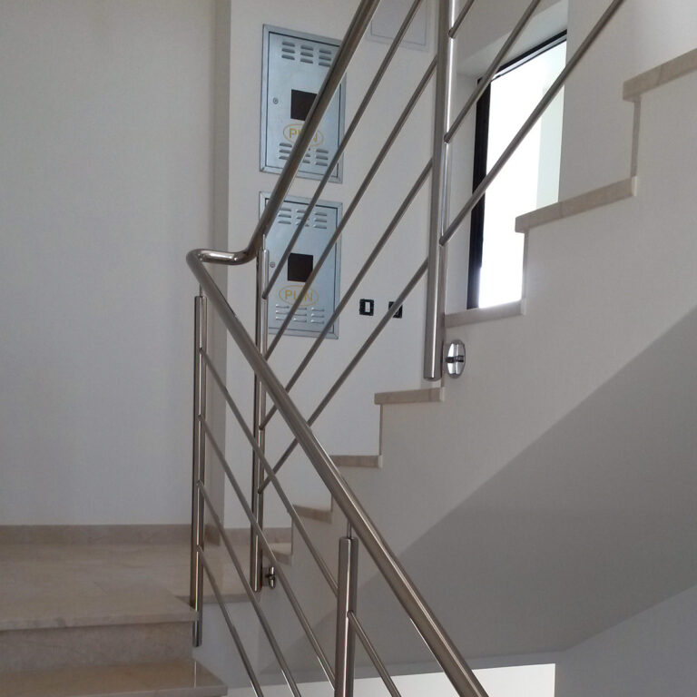 Moderno unutarnje stubište sa svijetlim stepenicama i elegantnom ogradom od inoxa. Stubište vodi prema gore, s električnim ili komunalnim razvodnim pločama na zidu u pozadini. Prirodno svjetlo struji kroz prozor.