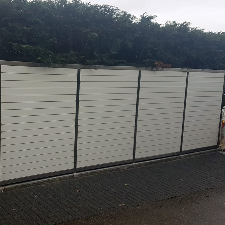 Bijela horizontalna ograda od letvica s metalnim stupovima stoji ispred gustog zelenog grmlja po oblačnom danu; tlo je mokro, što ukazuje na nedavnu kišu - savršeno za predstavljanje ograde od inoxa i mesinga.