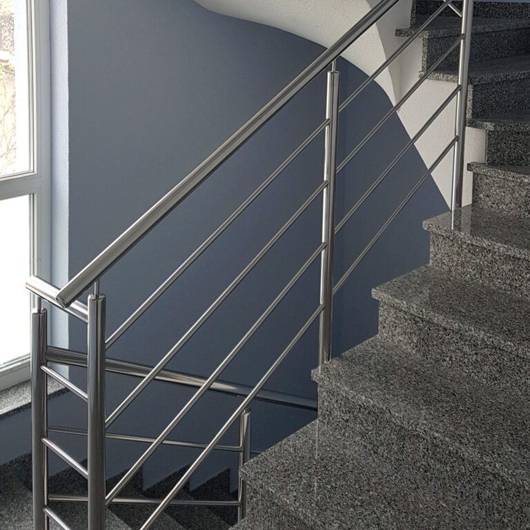 Pogled izbliza na moderno unutarnje stubište s poliranim sivim kamenim stepenicama i Unutarnje ograde od inoxa, pored velikog prozora koji propušta prirodno svjetlo.