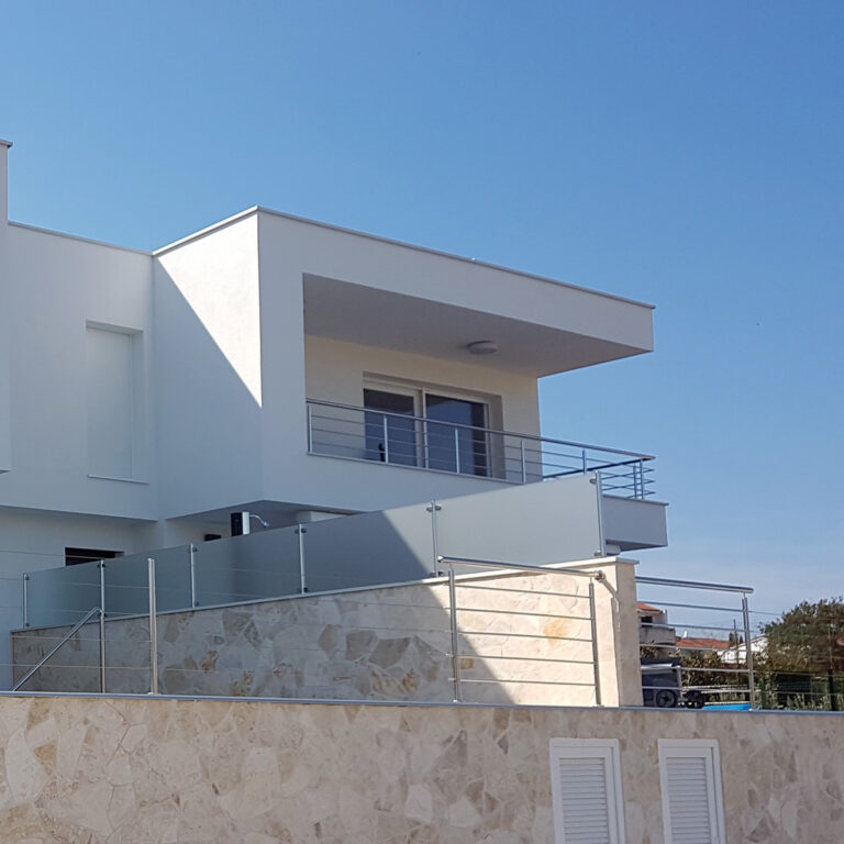 Moderna bijela kuća s čistim linijama i ravnim krovom, s velikim prozorima i balkonom koji obuhvaća cijelu kuću, smještena uz vedro plavo nebo. Donji dio ima kamenu fasadu i elegantne ograde od inoxa i mesinga sa staklenim ogradama.