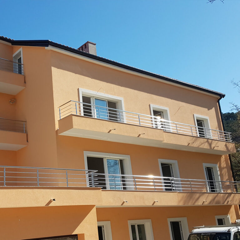 Moderna, svijetlonarančasta trokatnica s više balkona s ogradama od inoxa i mesinga, velikim prozorima i vedrim plavim nebom u pozadini.