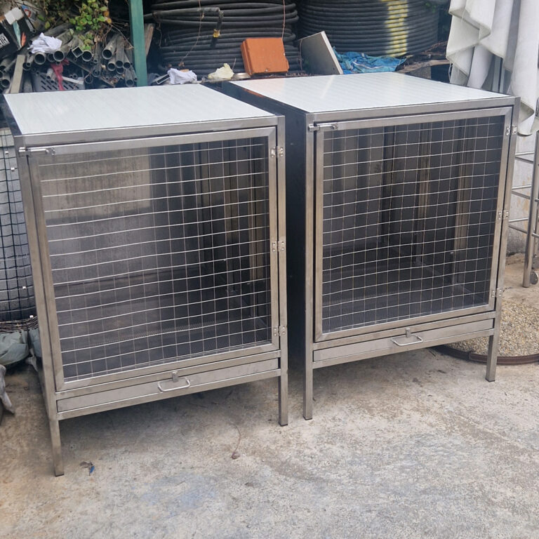Dva inox kaveza od nehrđajućeg čelika s vratima od žičane mreže i malim ladicama na dnu stoje jedan pored drugog na betonskoj površini, okruženi raznim industrijskim materijalima u pozadini.