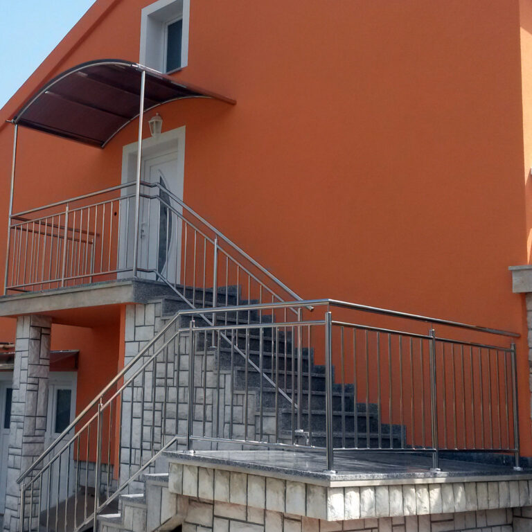 Vanjsko stubište s ogradama od inoxa i mesinga i kamenim stepenicama koje vode do bijelih vrata na narančastoj zgradi; odmorište ima smeđu zakrivljenu nadstrešnicu iznad vrata.