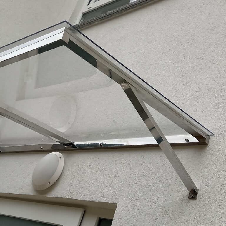 Pogled izbliza odozdo na stakleno-metalnu tendu, ili nadstrešnice od inoxa, postavljenu iznad bijelih vrata na svijetloj zgradi, s okruglim vanjskim svjetlom na zidu ispod nje.