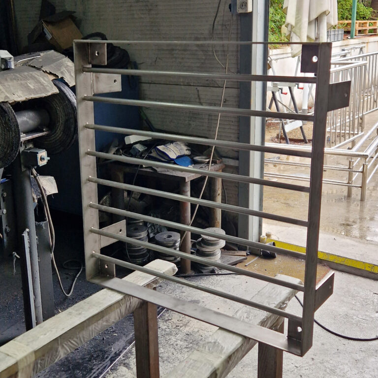 Pravokutni metalni okvir s horizontalnim šipkama, koji nalikuje malim vratima ili rešetki od inox galanterije, stoji uspravno u industrijskoj radionici s alatima, opremom i metalnim dijelovima u pozadini.