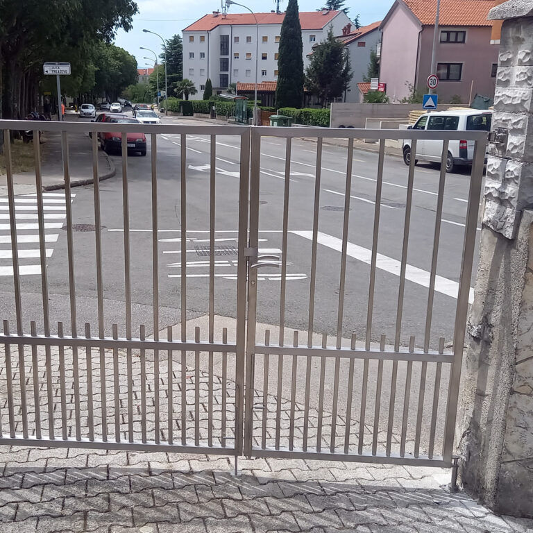 Nehrđajući čelik blokira pločnik, okrenut prema ulici s automobilima i kućama. Zatvorena vrata nalaze se uz pješački prijelaz i prometne oznake, s drvećem i zgradama u pozadini.