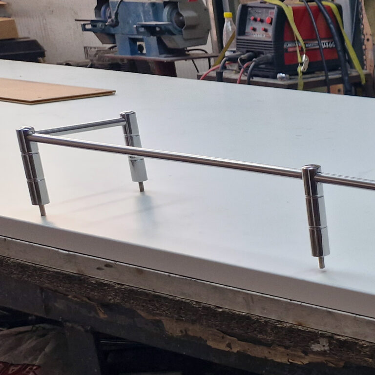 Metalna ručka od inox galanterije s cilindričnim nosačima leži na bijeloj površini stola u radioničkom okruženju, okružena alatima i opremom u pozadini.