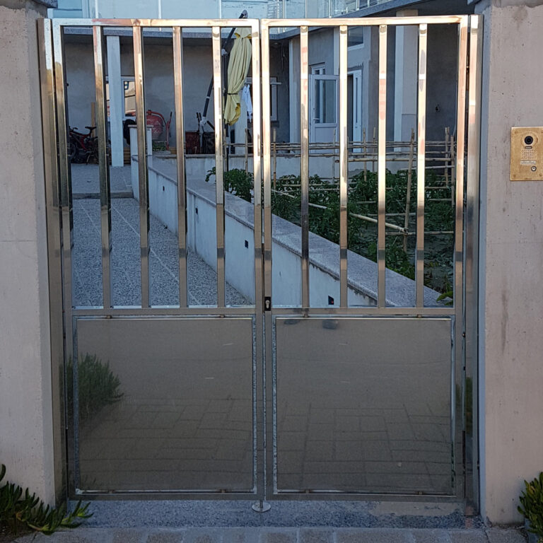 Sjajni portuni od inoxa s okomitim šipkama i donjom punom pločom stoji između dva betonska stupa, vodeći u dvorište s pješačkom stazom, biljkama i nekoliko zgrada u pozadini.