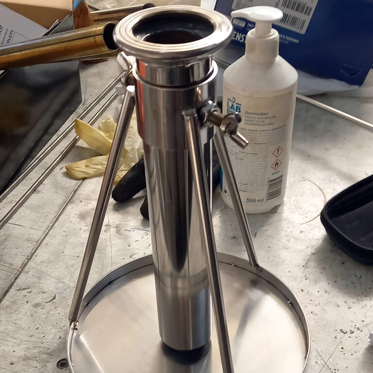 Sjajni metalni cilindrični instrument od inox galanterije s tri potporne šipke na okruglom postolju stoji na zatrpanoj radnoj površini, okružen bijelom bocom, rukavicama i raznim laboratorijskim predmetima u pozadini.