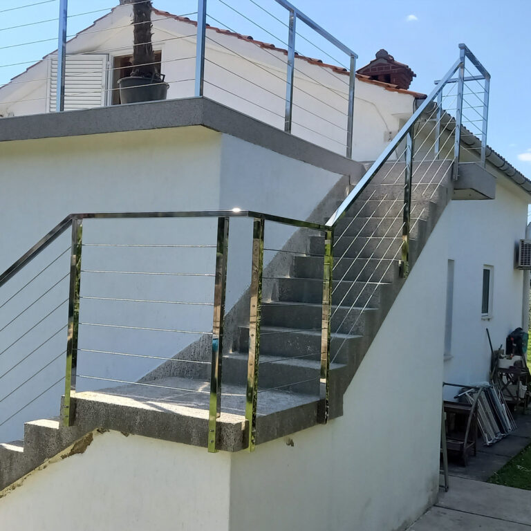 Vanjsko betonsko stubište s rukohvatima od inoxa i mesinga vodi na gornji kat bijele zgrade. Stepenice su omeđene popločanim dijelom terase, zgrada ima crijepni krov, a ispod je vidljiv i namještaj.