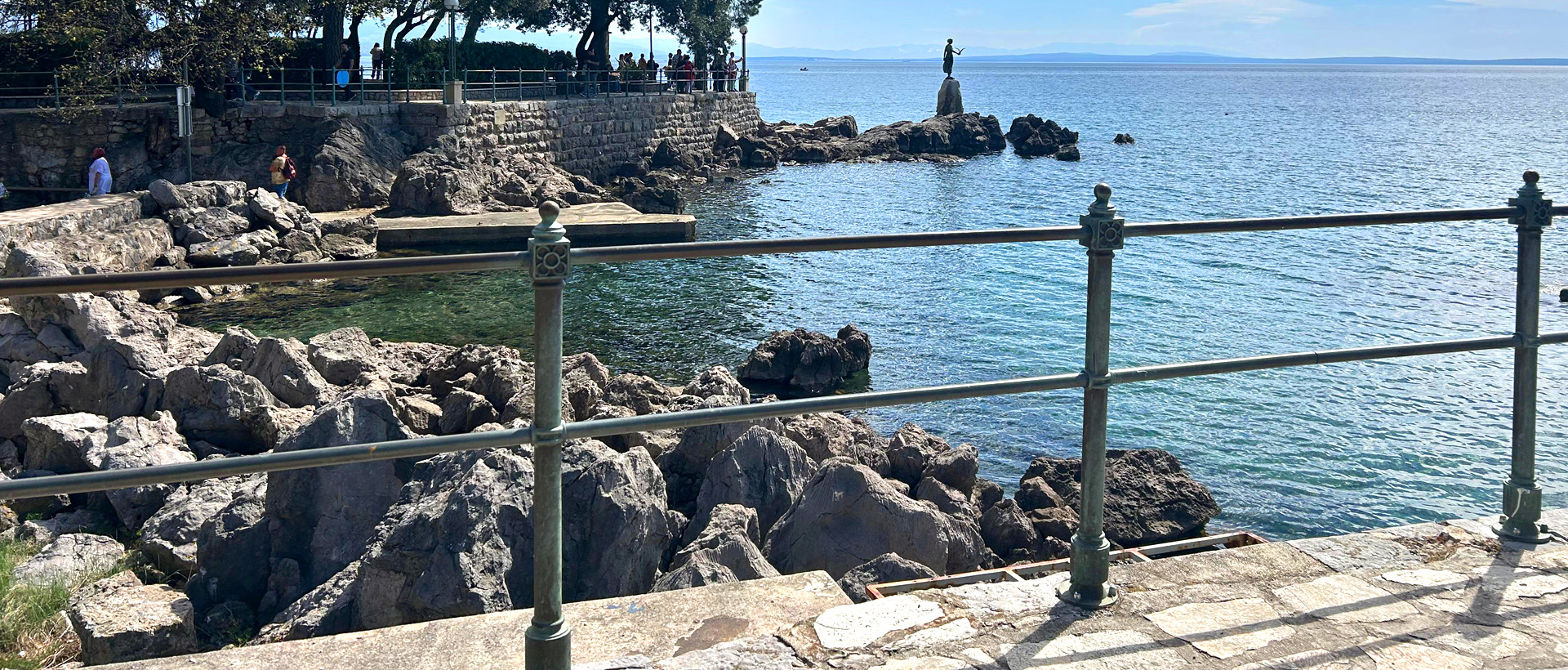 Pogled na stjenovitu obalu s bistrom plavom vodom, kip na stijeni i ograde od inoxa u prvom planu. Ljudi su vidljivi blizu kamenog zida, a drveće pruža hlad s lijeve strane.