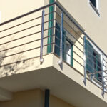 Kutni balkon s metalnom ogradom pričvršćen za bež zgradu, s prozorima sa zelenim okvirima i vanjskim kapcima. Sunčeva svjetlost stvara oštre sjene na zidu.