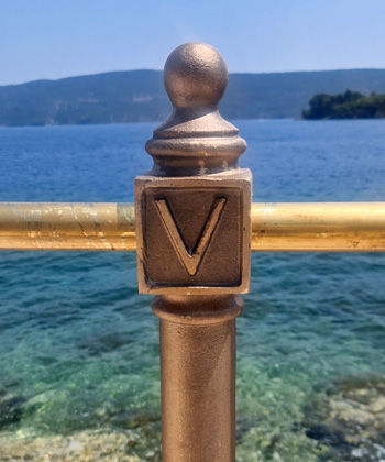 Brončani stup s uzdignutim slovom V stoji blizu ograde od inoxa ispred čistog plavog mora, s udaljenim brdima i trakom stjenovite obale vidljivim u pozadini pod sunčanim nebom.
